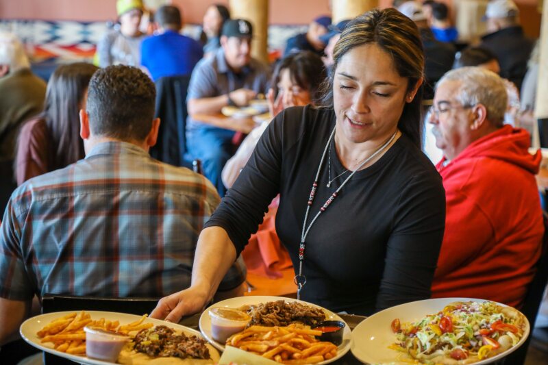 A server at Indian Pueblo Kitchen delivers three plates of enchiladas and more, with seated diners in background