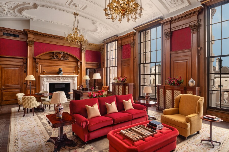 Interior of Haldane Suite at the Raffles London at the OWO, with dark wood paneling, a red sofa, floor-to-ceiling windows, and gilt chandeliers