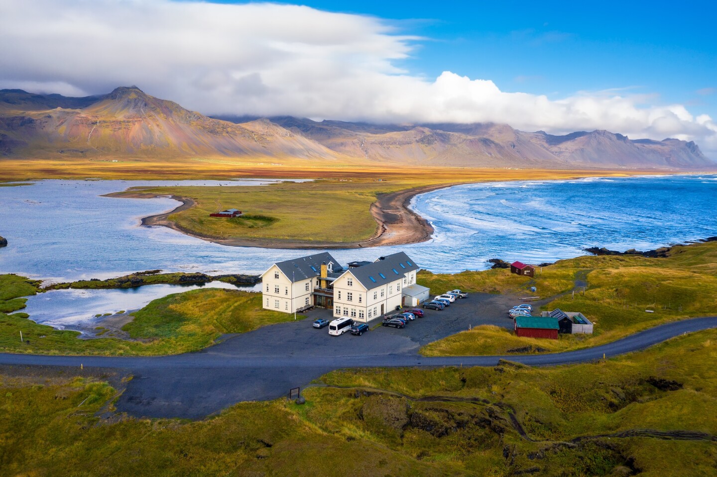 A hotel with cars in a small parking lot on a paved road next to a coastline with cloud-covered mountains in the background