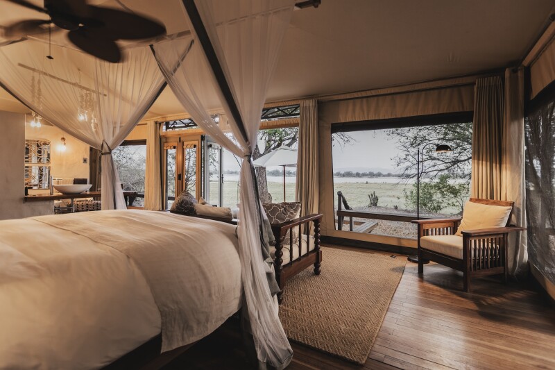 Beige guest room at Wilderness Chikwenya Camp, with mosquito netting around bed and large windows overlooking surrounding landscape