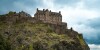 A castle atop a green rocky hill, with clouds overhead