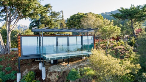 A glass house nestled on a cliff in Big Sur, surounded by trees and shrubs