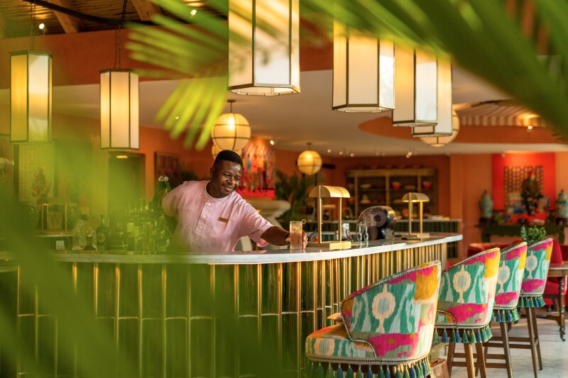 A bartender places a drink on the bar at Masiya's Camp, in a room with brightly colored chairs in turquoise and magenta hues.