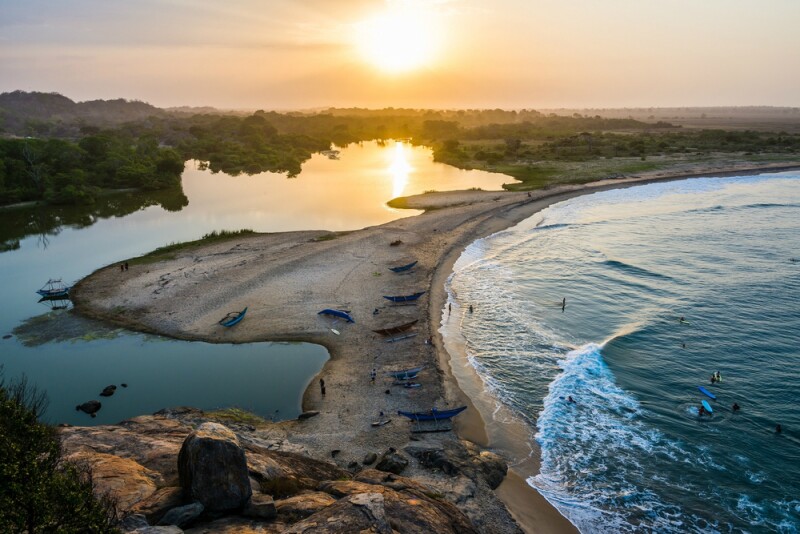 A curved beach with a thin strip of sand and a lagoon behind it, with a golden sunset low in the sky