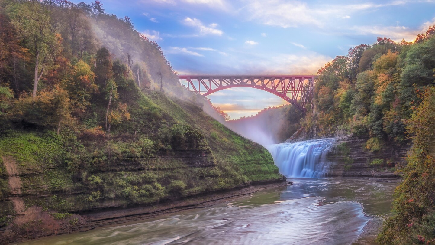 A river and waterfall flow along the striking limestone rock canyon of Letchworth State Park with a soaring red bridge in the background and trees in fall colors all around