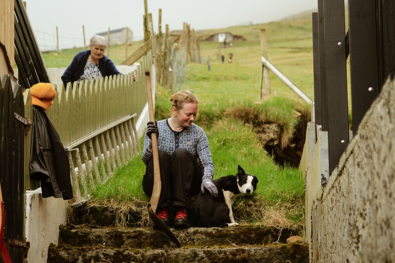 Woman sits on outdoor staircase holding a shovel and petting a black and white puppy while an elderly woman looks on from behind