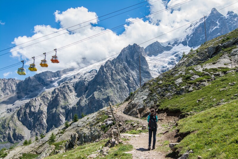 Person walking on narrow path, with jagged mountains and four small aerial trams in background