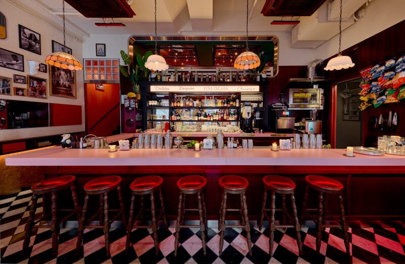 Interior of bar with tiled floor, row of stools, vintage ceiling lamps, and framed photographs on left walls