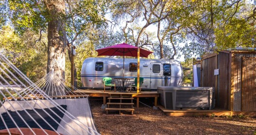 An Airstream trailer with a hot tub, outdoor shower, deck, and hammock, surrounded by trees