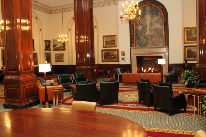 The high-ceilinged lobby of the Union League Club, with large, dark wood columns, chandeliers, framed art on walls, and lit fireplace